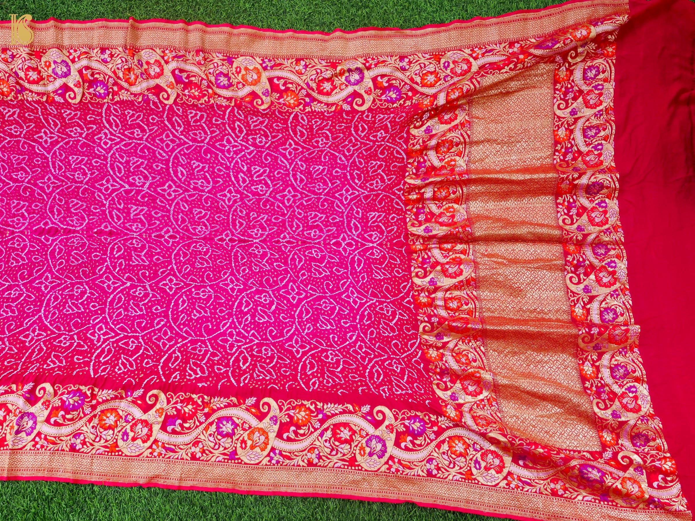 A bright pink saree or fabric piece with intricate white floral patterns and a wide decorative border in red, orange, and gold, laid flat on green grass.