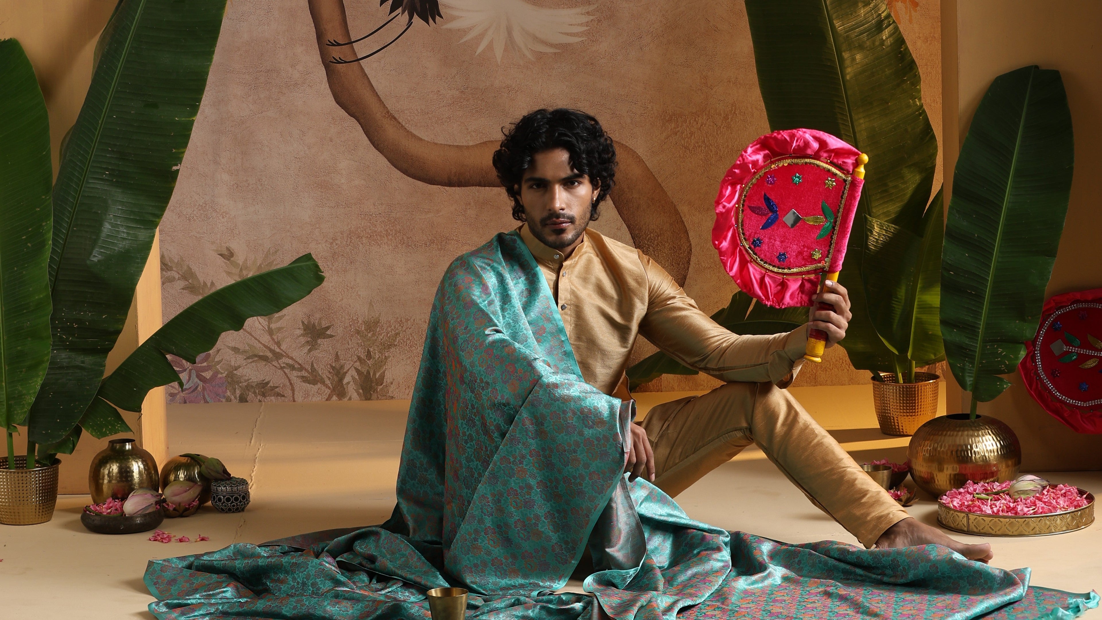 Man sitting on the floor holding a colorful fabric item with plants and decorative items in the background