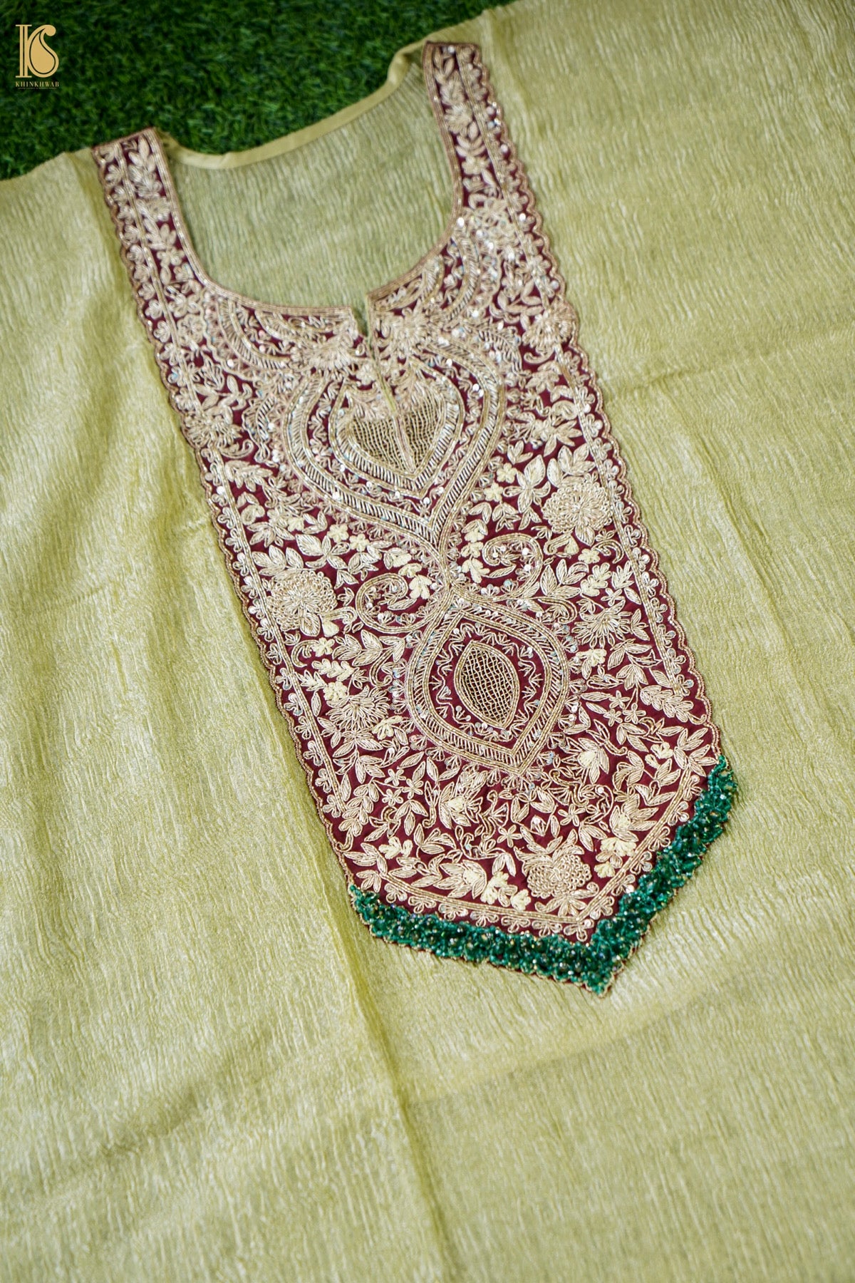 A close-up of a light olive green fabric garment laid flat, featuring a large embroidered yoke panel in beige and burgundy with a green beaded border.