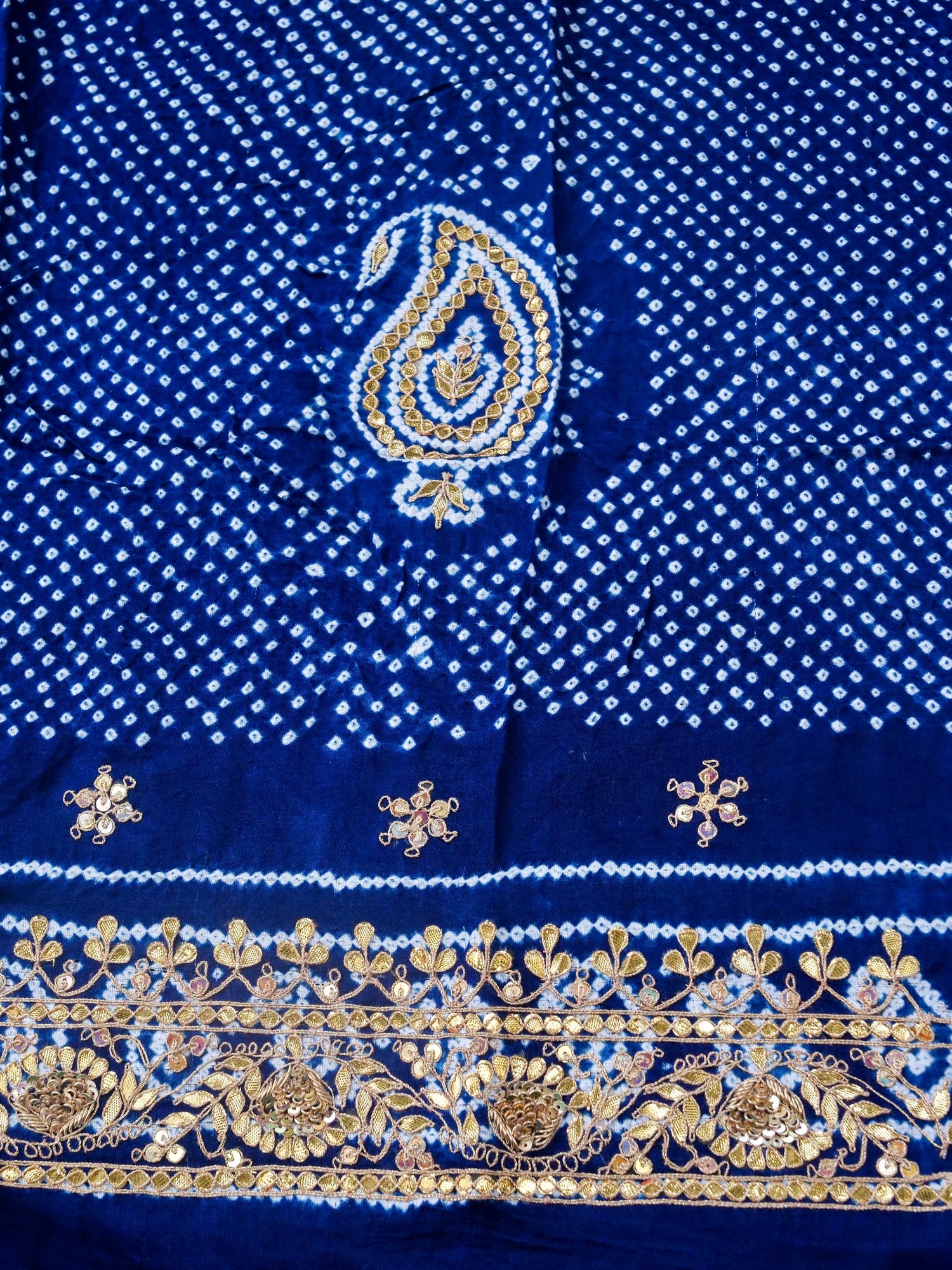 A close-up of a royal blue fabric with white bandhani dot patterns, a central paisley motif in blue and gold, and an ornate gold floral border along the bottom edge.