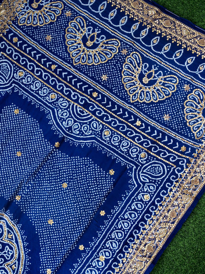 A close-up of a royal blue patterned textile laid on green grass, showing ornate gold and white paisley and floral motifs with dense dot work.