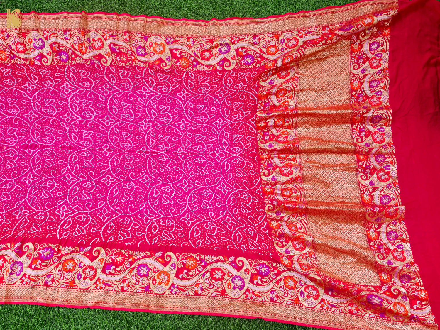 A bright pink saree or fabric piece with intricate white floral patterns and a wide decorative border in red, orange, and gold, laid flat on green grass.