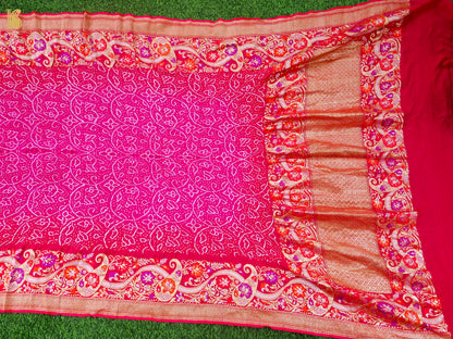 A bright pink saree or fabric piece with intricate white floral patterns and a wide decorative border in red, orange, and gold, laid flat on green grass.