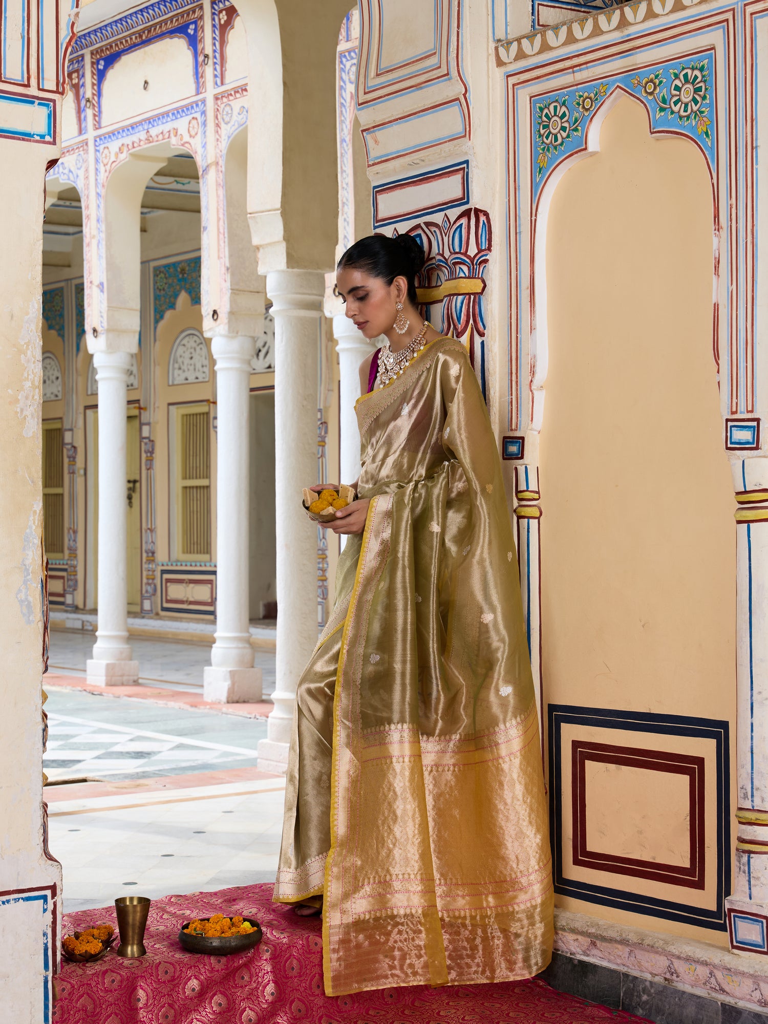 A woman wearing a shimmering gold saree stands in a traditional courtyard with arched walkways and ornate walls, holding a small bowl of offerings.