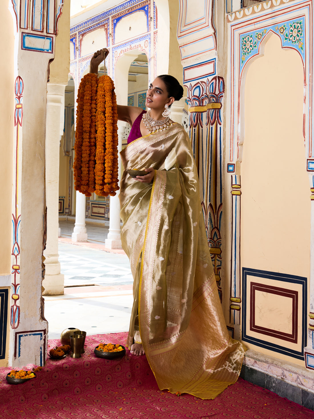A woman in a traditional Indian outfit wearing a shimmering gold saree and a purple blouse stands in an ornate palace corridor, holding a long orange marigold garland.