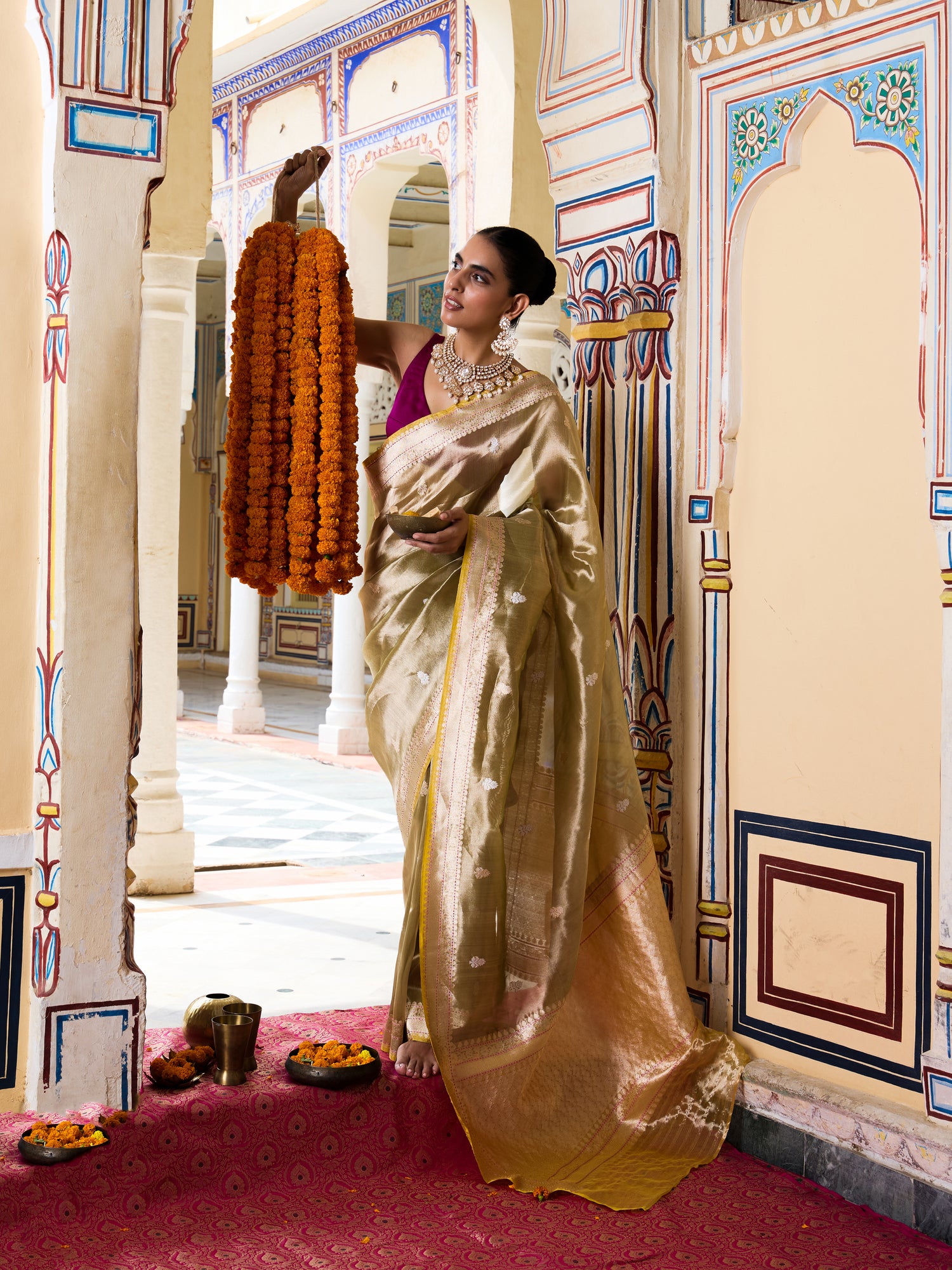 A woman in a traditional Indian outfit wearing a shimmering gold saree and a purple blouse stands in an ornate palace corridor, holding a long orange marigold garland.