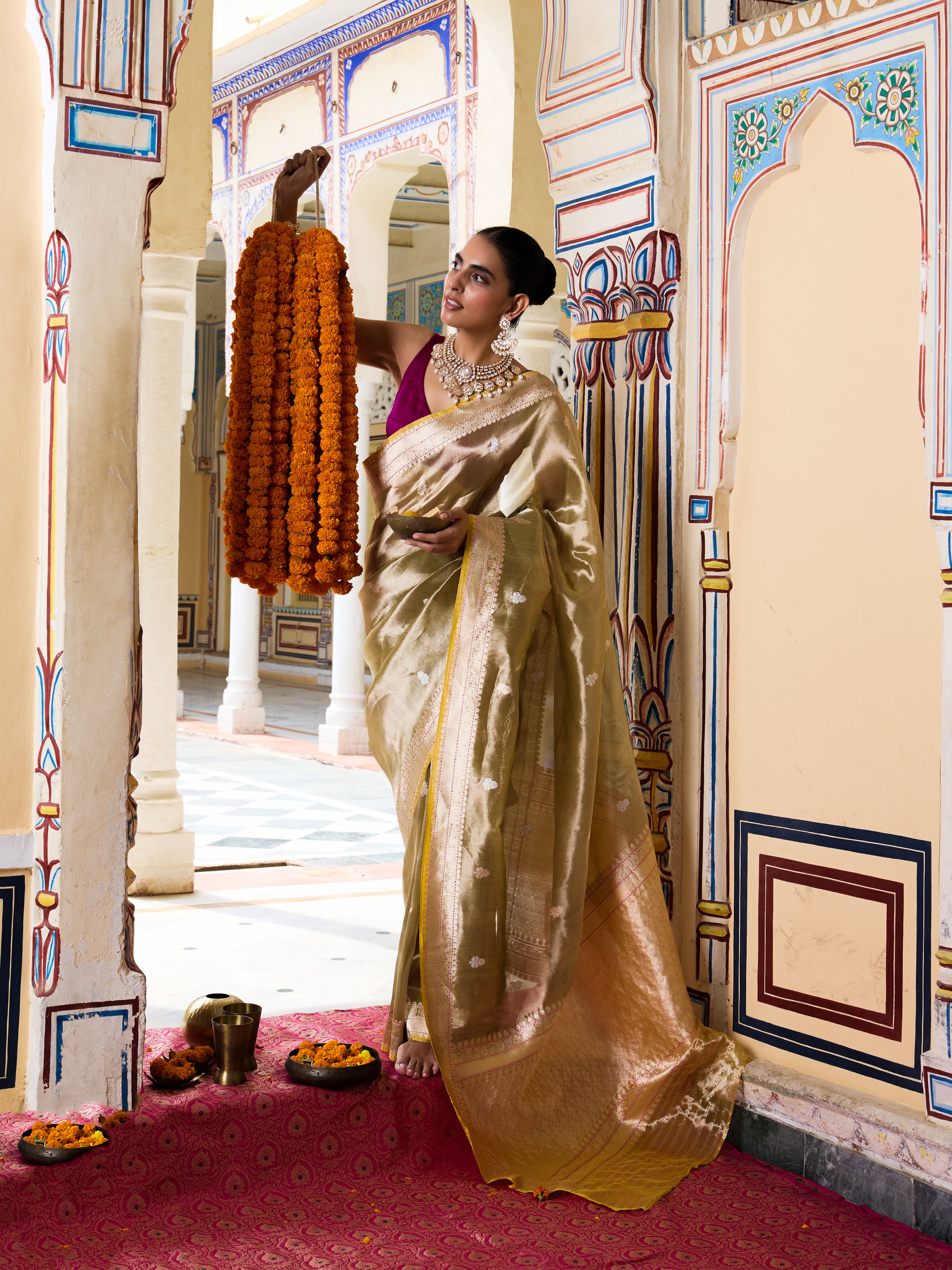 A woman in a traditional Indian outfit wearing a shimmering gold saree and a purple blouse stands in an ornate palace corridor, holding a long orange marigold garland.