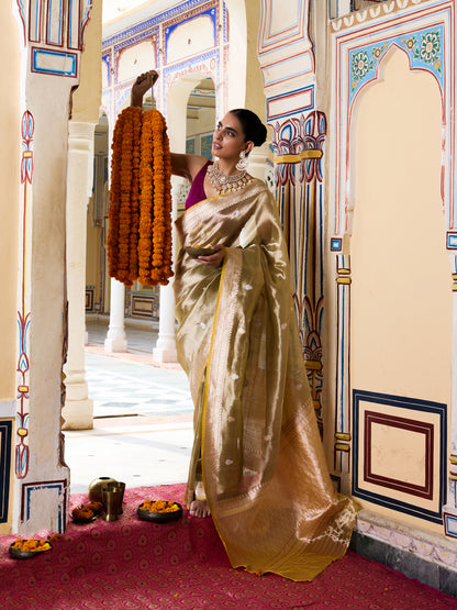 A woman in a traditional Indian outfit wearing a shimmering gold saree and a purple blouse stands in an ornate palace corridor, holding a long orange marigold garland.