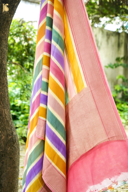 A close-up of a draped multicolored striped fabric, likely a saree, hanging outdoors next to a tree trunk with a blurred garden background.