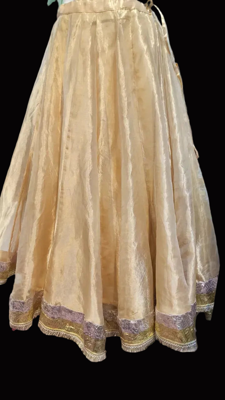 A voluminous gold-colored traditional skirt with a shimmering satin-like finish and a decorative embroidered border, displayed indoors on a patterned rug.