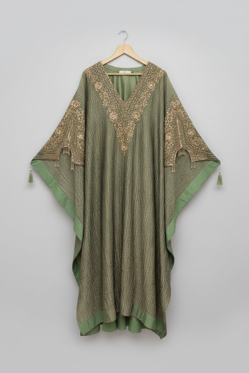 An olive green kaftan dress with intricate gold embroidery and beadwork, hanging on a wooden hanger in front of an ornate carved wooden mirror frame.