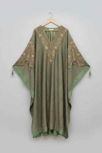 An olive green kaftan dress with intricate gold embroidery and beadwork, hanging on a wooden hanger in front of an ornate carved wooden mirror frame.