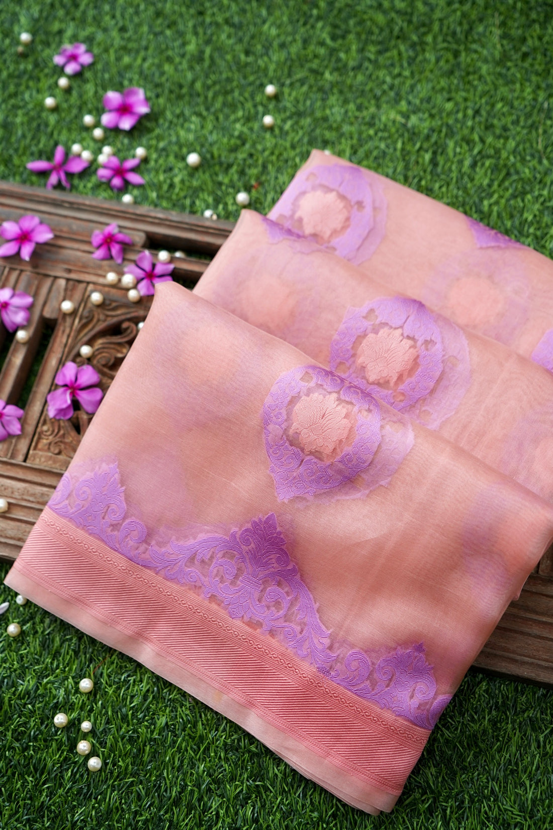 A peach-pink and purple saree with floral motifs and a decorative border, draped on a wooden tray over a green grass background with small purple flowers.