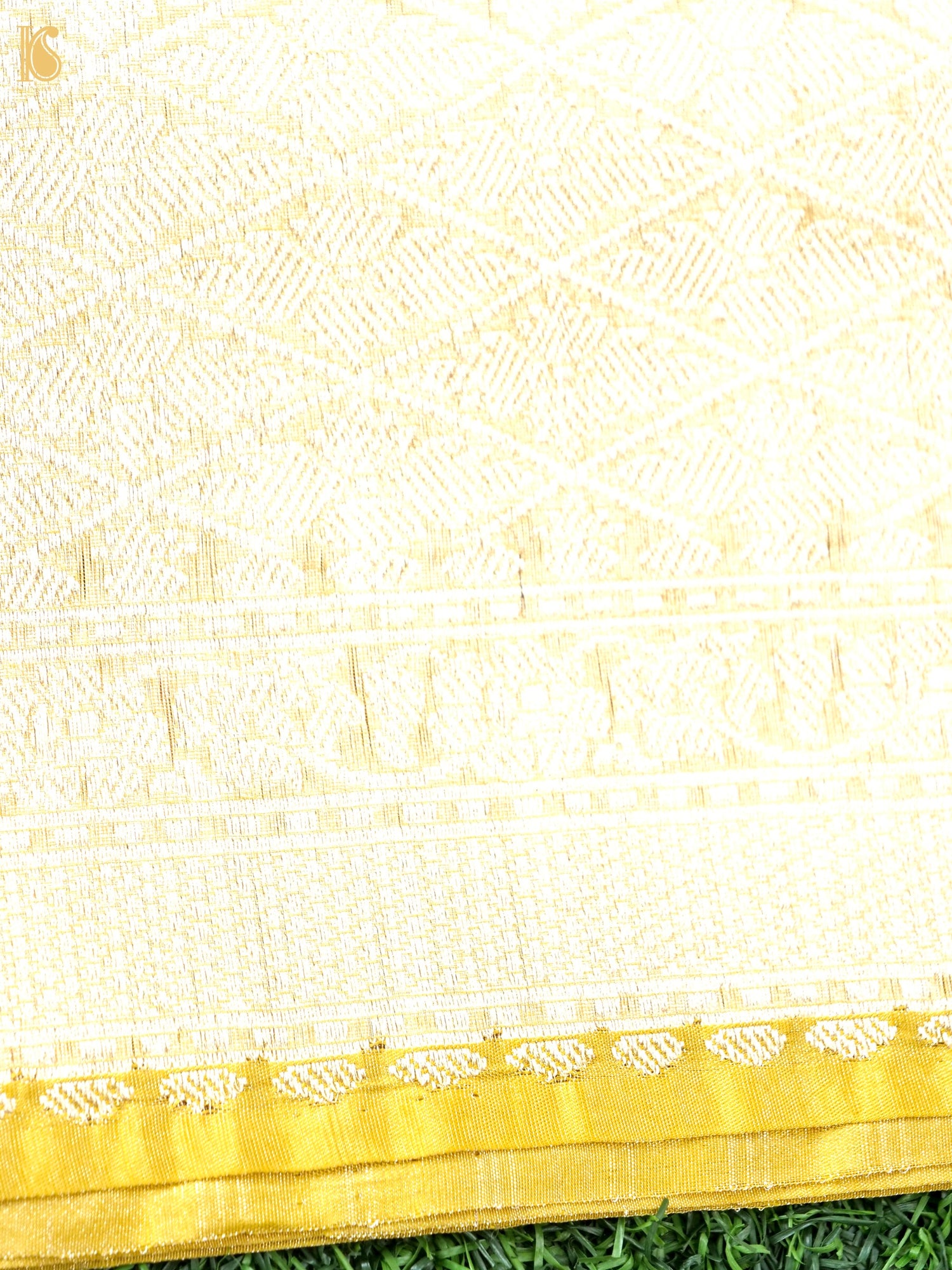 A close-up of a cream or off-white woven textile with a gold border and subtle woven geometric and floral patterns, laid over green grass.