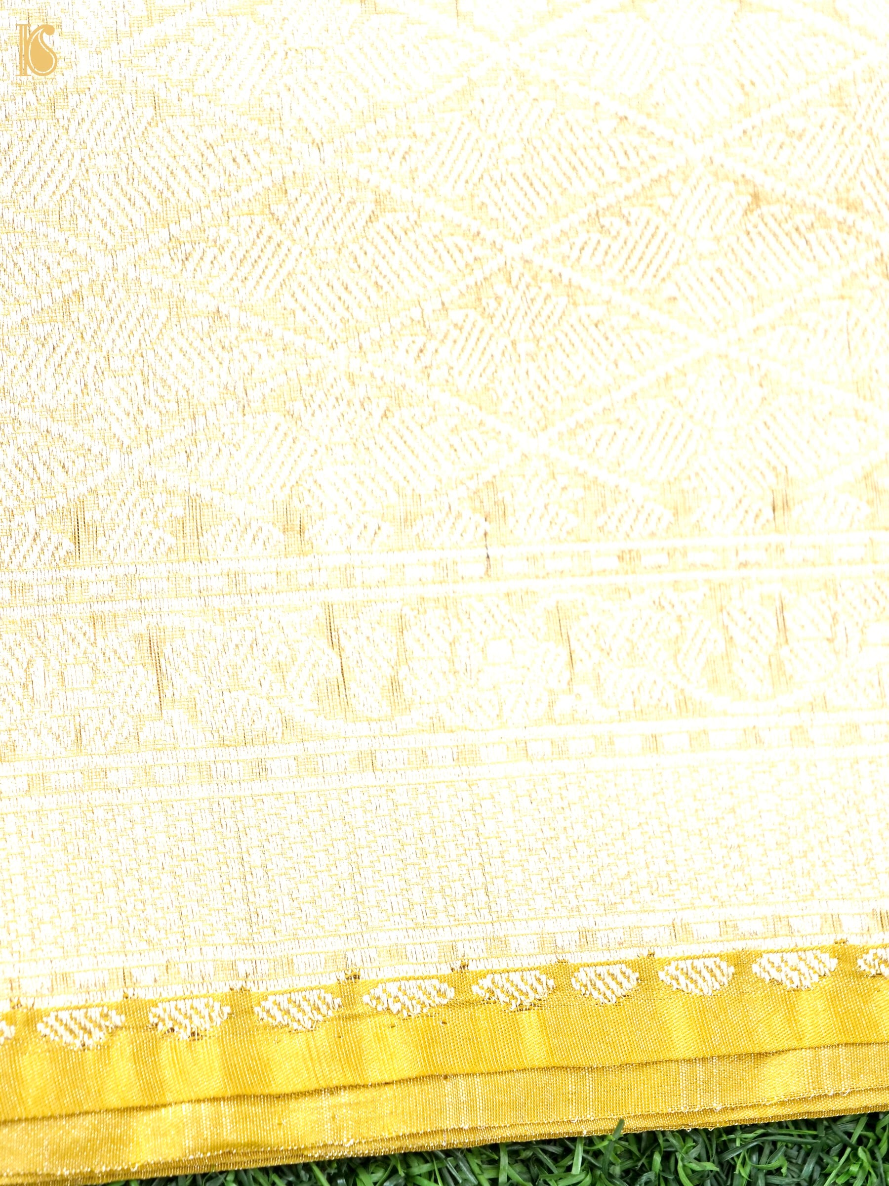 A close-up of a cream or off-white woven textile with a gold border and subtle woven geometric and floral patterns, laid over green grass.