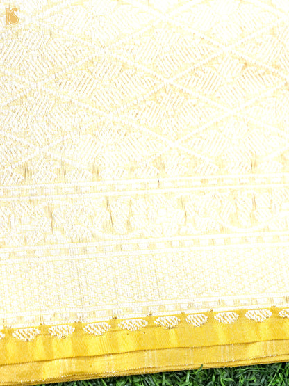 A close-up of a cream or off-white woven textile with a gold border and subtle woven geometric and floral patterns, laid over green grass.