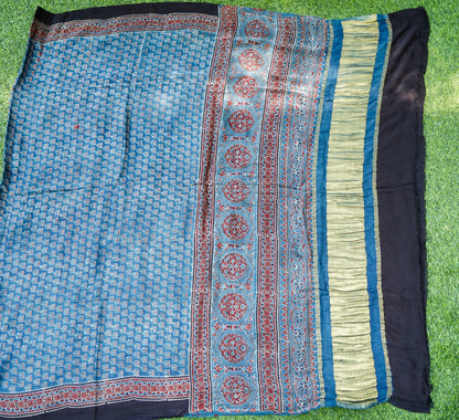 A blue patterned fabric, likely a saree or scarf, laid flat on green grass. The fabric features a light blue body with a repeating geometric pattern, a central border with red and gold circular motifs, and a wide black border on the right side.