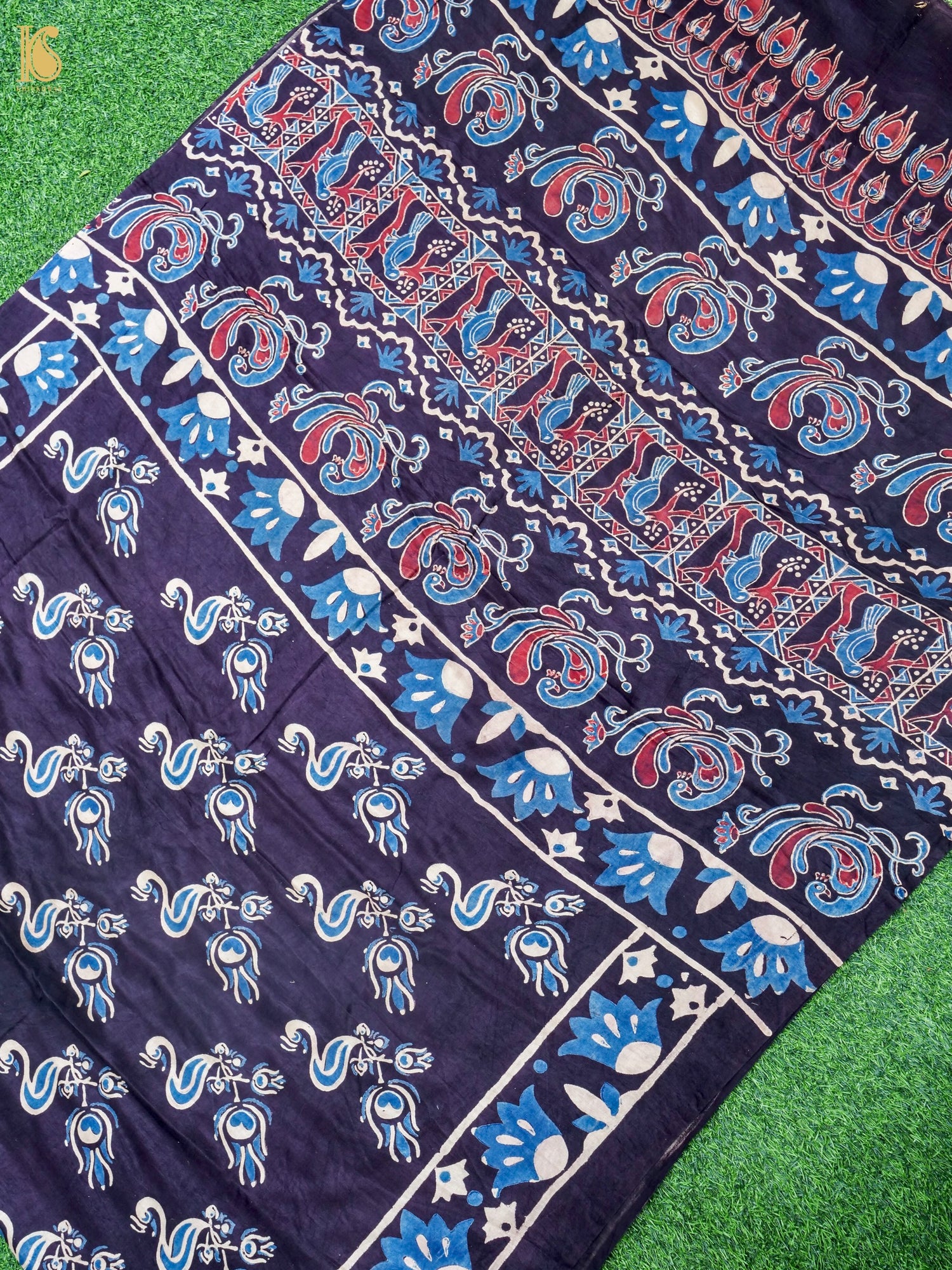 A close-up of a dark navy blue fabric with intricate hand-block printed patterns in blue, red, and white, laid flat on a green grass-like surface.