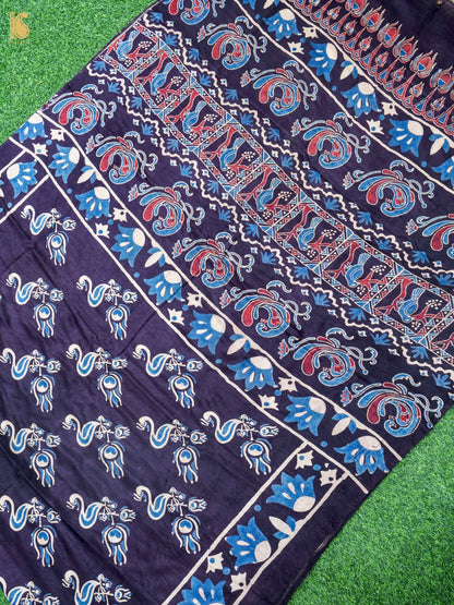A close-up of a dark navy blue fabric with intricate hand-block printed patterns in blue, red, and white, laid flat on a green grass-like surface.